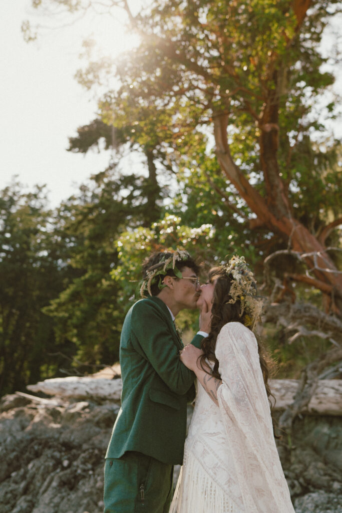 A documentary style wedding photo of a bride and groom kissing during golden hour in front of trees wearing flower crowns.