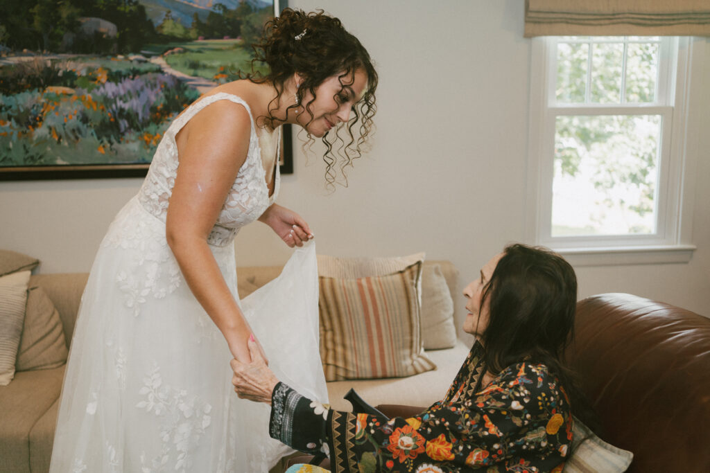 A documentary style wedding photograph of a bride holding hands with her grandmother who is sitting on a brown couch, wearing a silk floral robe.