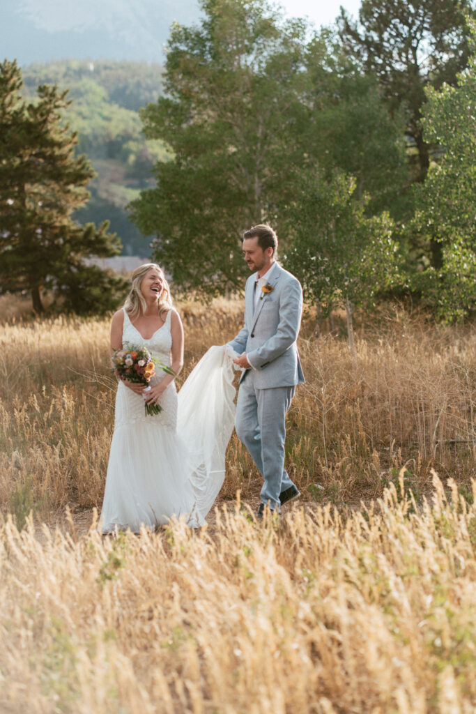 A groom holds the train and his bride walks in front of him, laughing candidly taken by a documentary style wedding photographer.