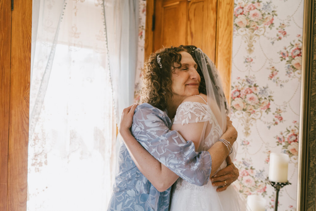 Documentary style wedding photography of a bride's mother embracing her in a hug with a soft smile on her face and her eyes closed.